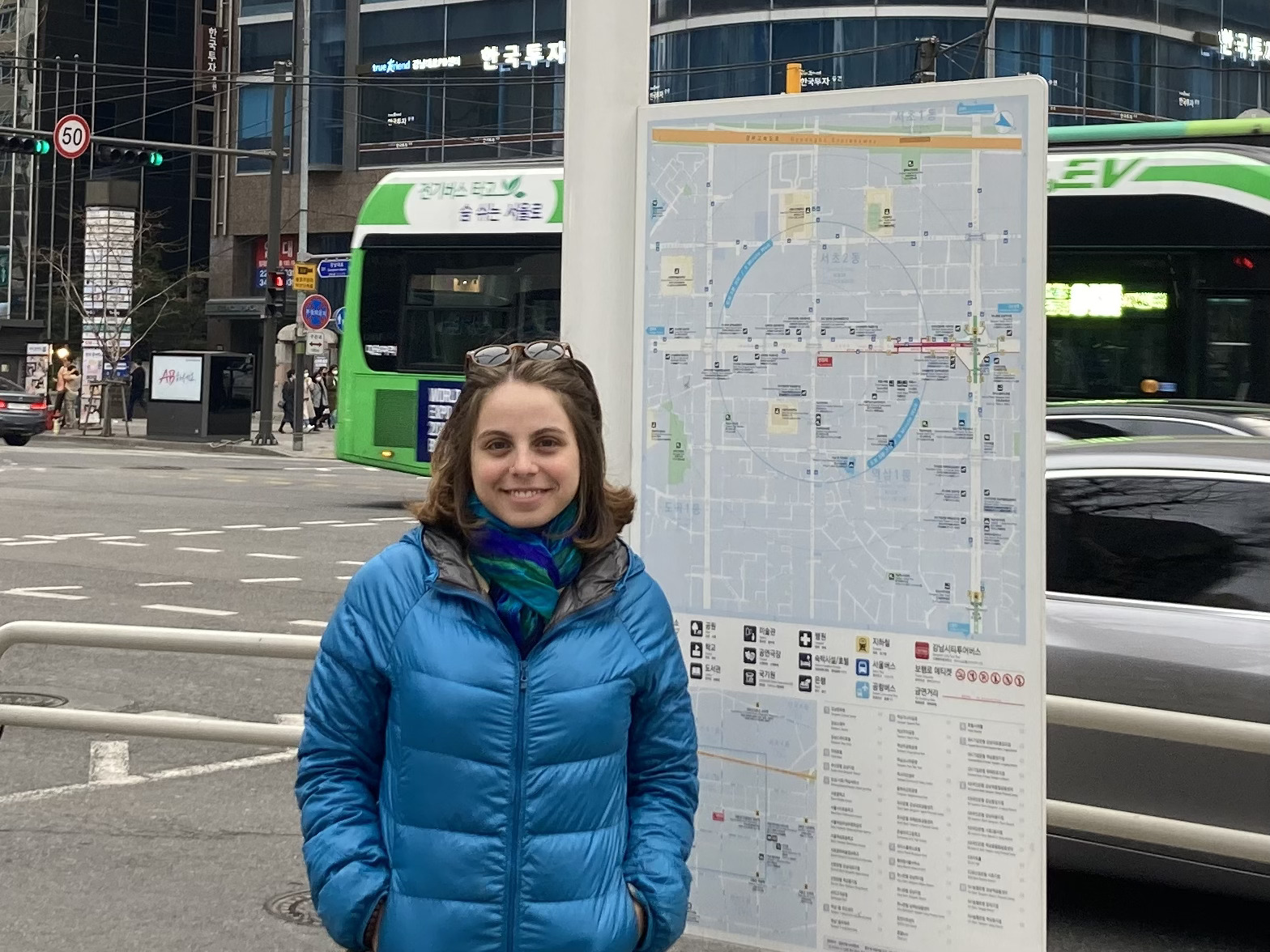 Sophie Arnstein wearing glasses on her head, a blue coat, and a blue-green scarf standing on the street in front of a map and smiling.