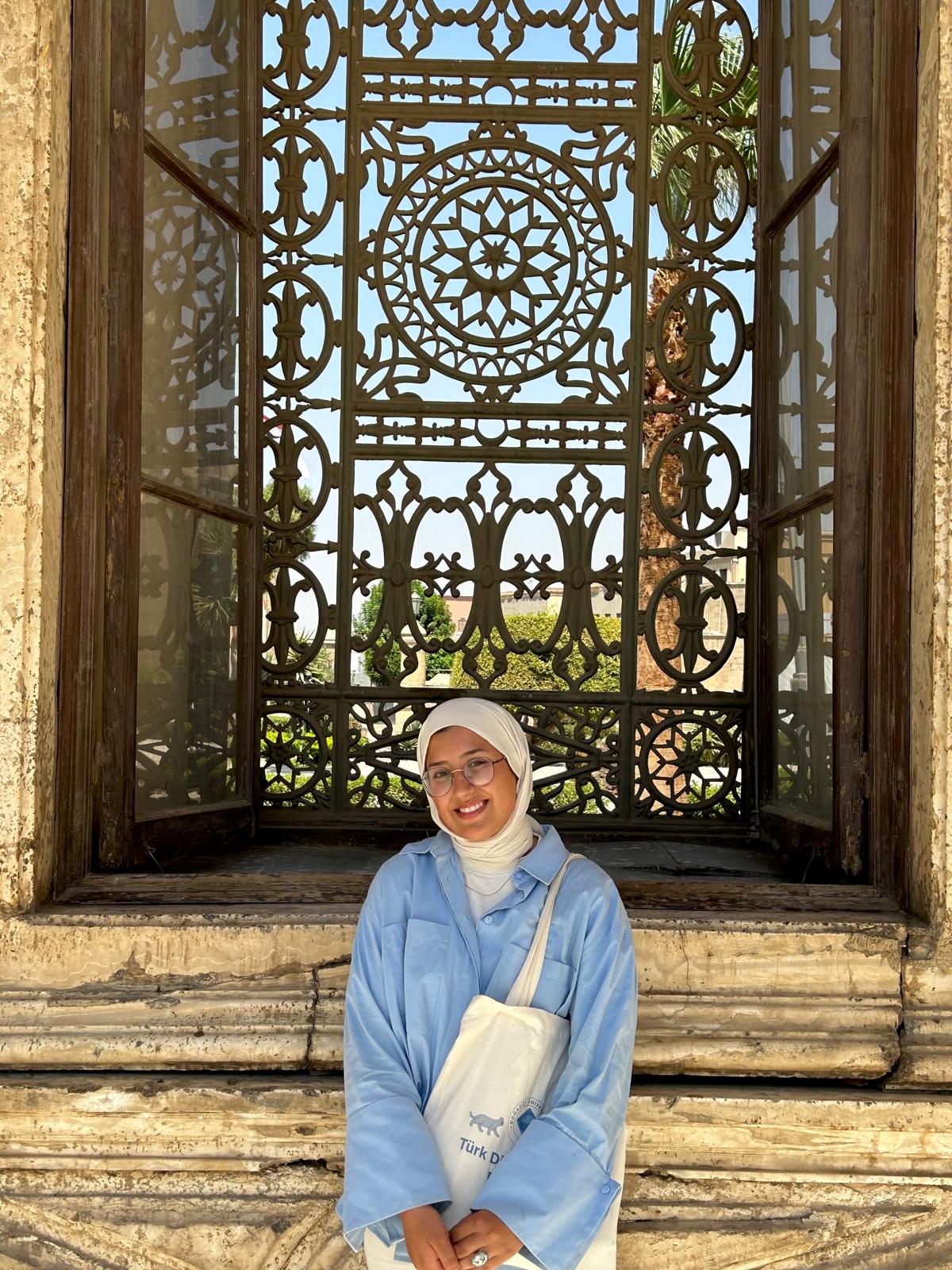 Aseel stands in front of an intricately patterned window with a smile on her face. She is wearing a periwinkle long-sleeve button down over a white garment, round wire-frame glasses, a white hijab, a thin chain, and a white tote bag. Her sleeve/arm mostly covers the tote bag, but "Türk D" (the rest of the word starting with "D" is covered) with a small animal cartoon above is visible.